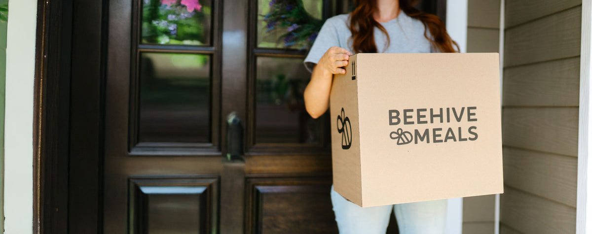 Women wearing a blue shirt holding a 'Beehive Meals' box in front of a door
