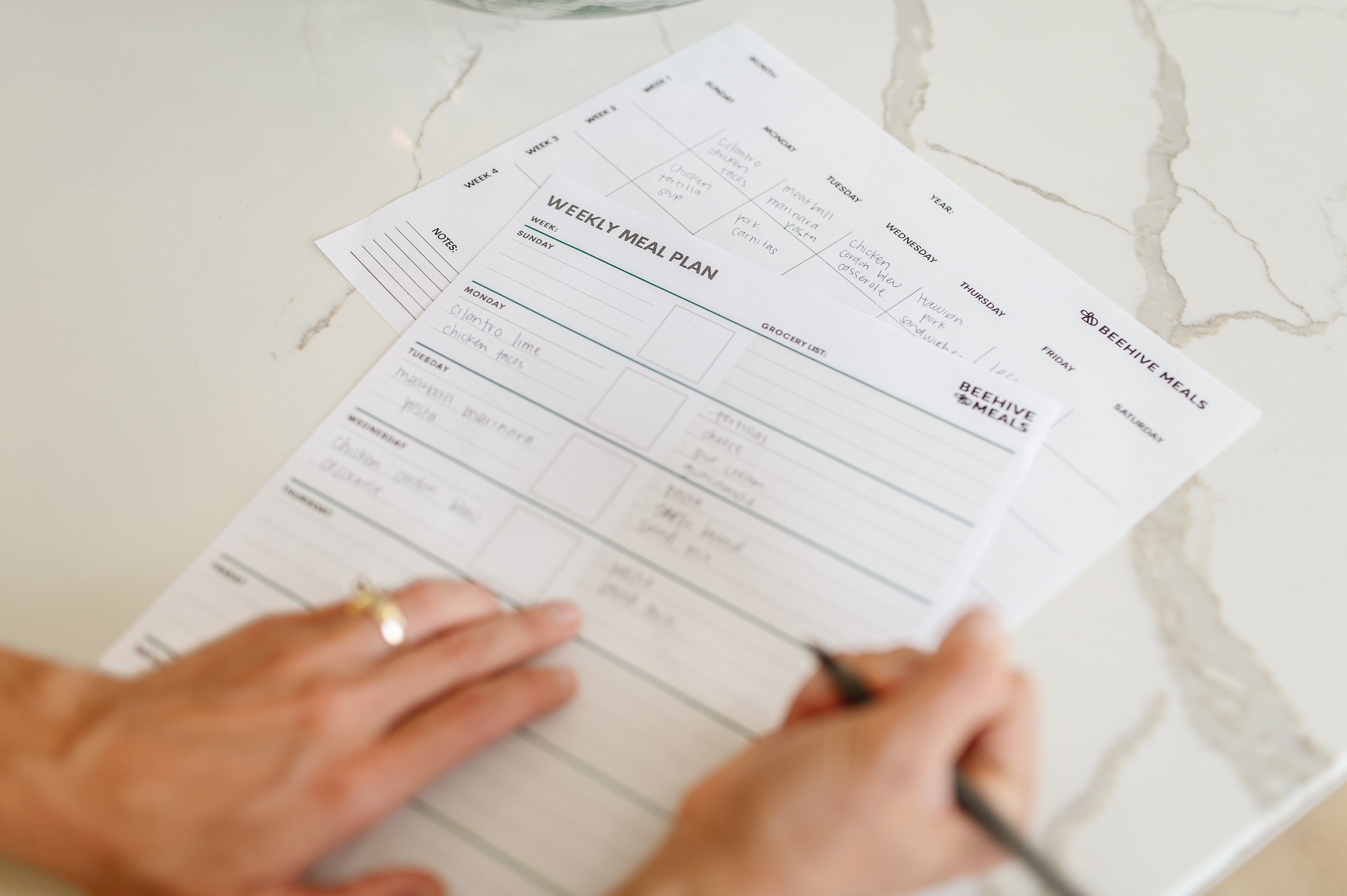 A person's hands are shown writing on white printable weekly meal plan sheets with black text, placed on a light marble surface.