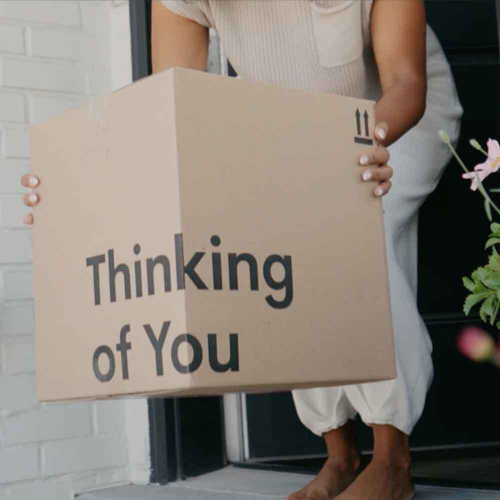 Person holding a cardboard box with 'Thinking of You' text, standing in front of a door.