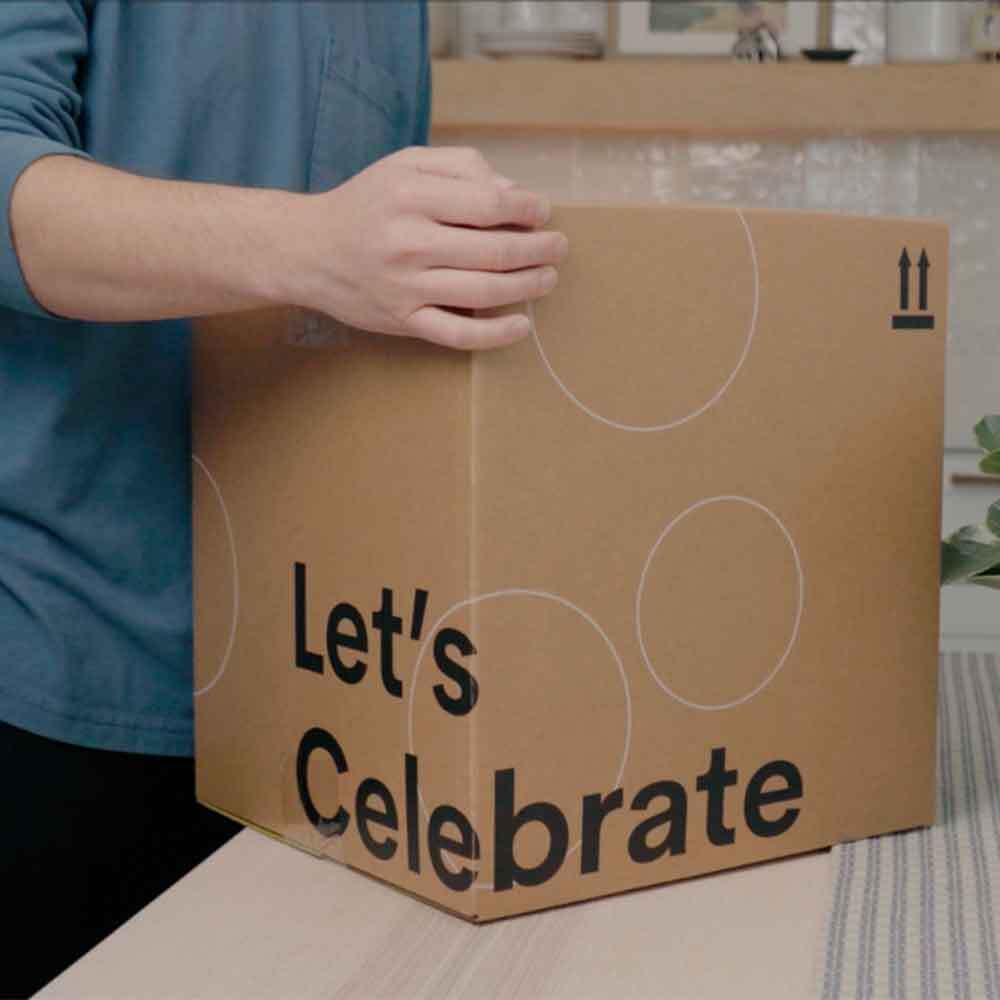 Person holding a cardboard box with 'Let's Celebrate' text in a kitchen setting