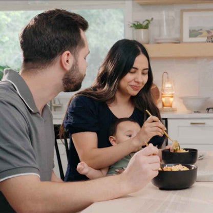 Family of three in a kitchen, with a woman holding a baby and a man sitting next to them.