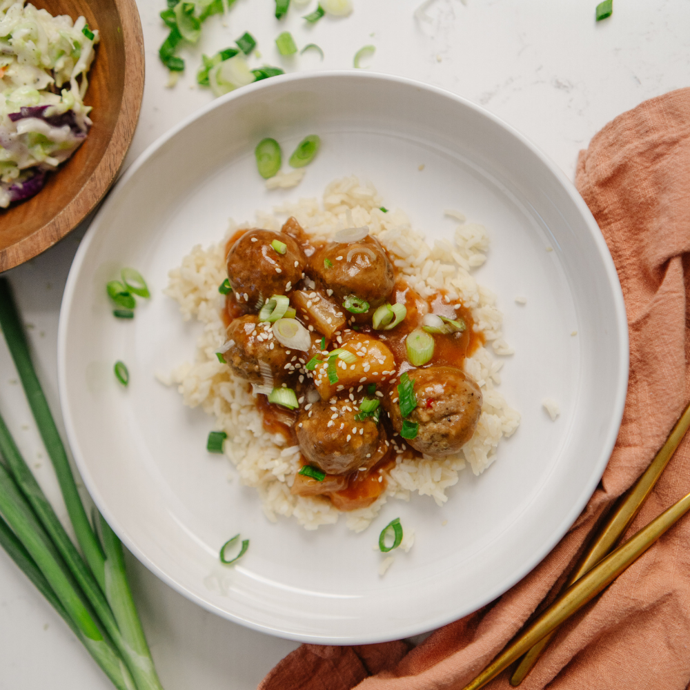 White plate with meatballs in a sauce over rice, garnished with green onions, on a light surface.