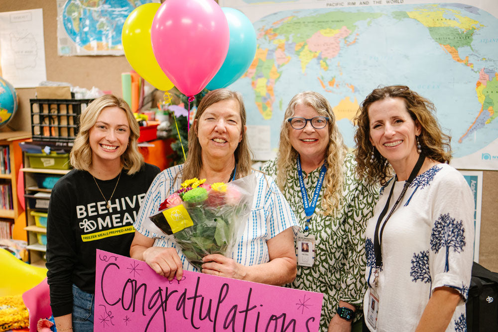 Four women standing together in a classroom or office setting, smiling and holding a pink 'Congratulations' sign, celebrating winning Beehive Meals. Balloons in pink, yellow, and teal float above them, and a world map is visible in the background.