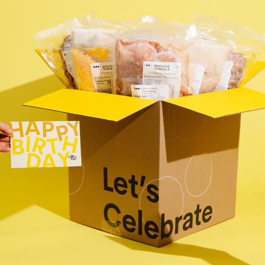 An open brown cardboard gift box with yellow flaps and the words 'Let's Celebrate' printed on the front, filled with packaged food items. A hand at the left edge holds a small card that says 'HAPPY BIRTH DAY' in yellow text.