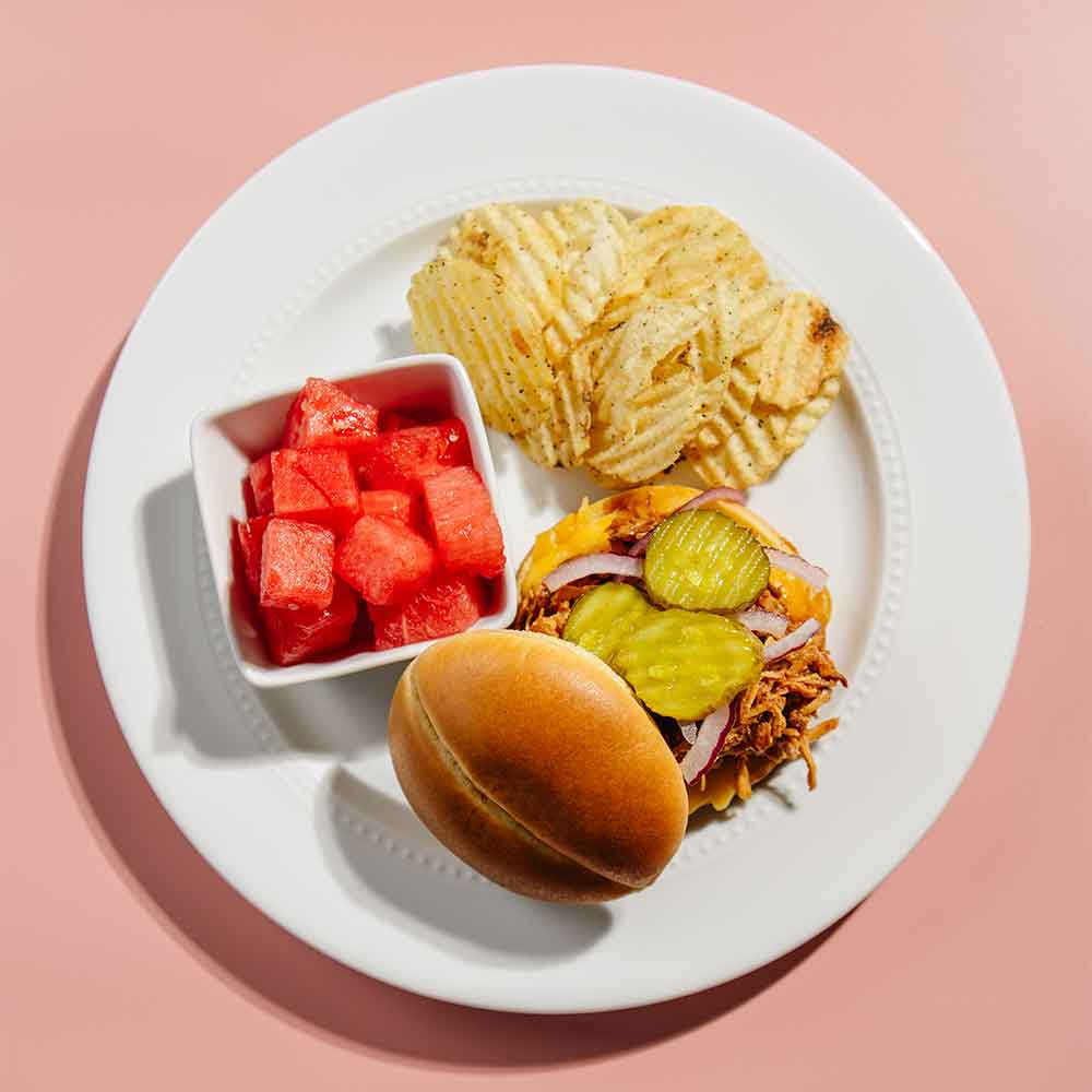 Plate with Beehive Meals BBQ chicken served on a bun with a side of watermelon, potato chips, and pickles