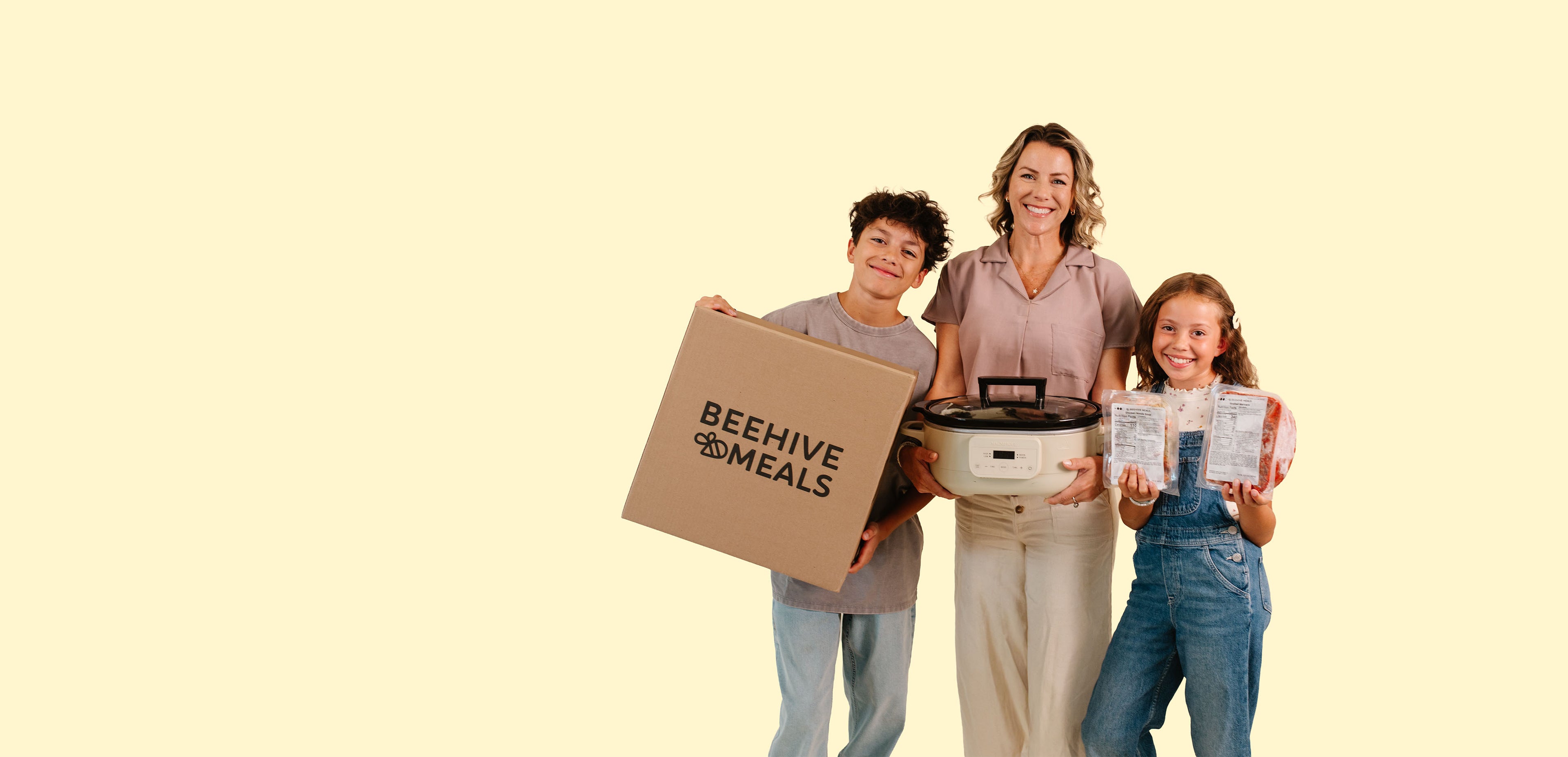 A family of three stands against a solid pale yellow background, holding products from Beehive Meals: a large cardboard box, a slow cooker, and frozen Beehive Meals.