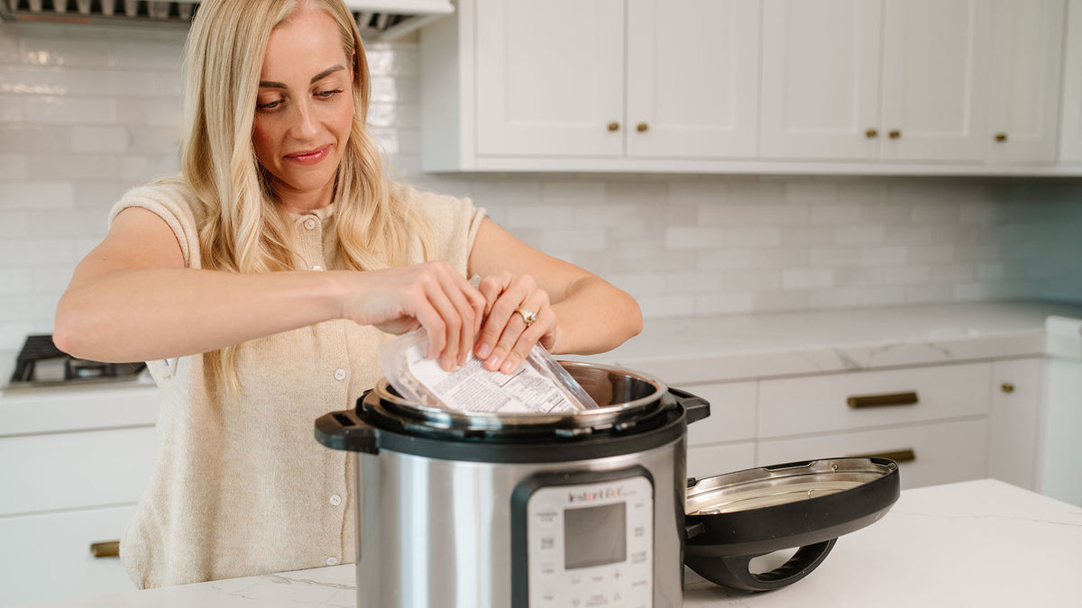 A woman in a beige top is shown from the waist up in a bright kitchen, placing a Beehive Meal into an Instant Pot pressure cooker. The Instant Pot is silver and black, and its lid is resting on the counter to the right.