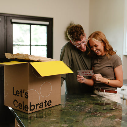 Two people in a kitchen with a 'Let's Celebrate' box and a 'Happy Couple' card.