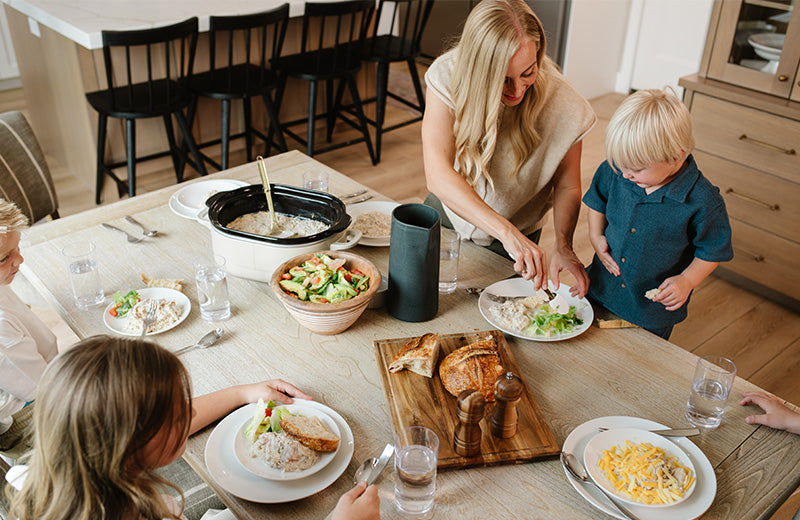 A family dining scene in a bright kitchen and dining area, with a woman serving food from a slow cooker at a wooden table while children eat plates of food. The scene includes a croissant on a cutting board, salad, pasta, and glasses of water.