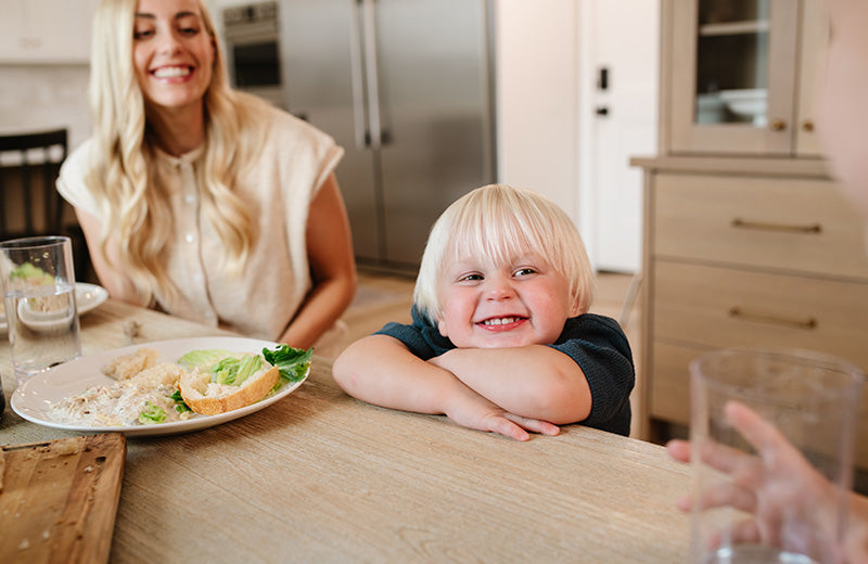 A smiling blonde toddler in a dark blue shirt leans on a wooden table in a bright kitchen, with a woman in a cream top seated in the background and a plate of food on the table in the foreground.