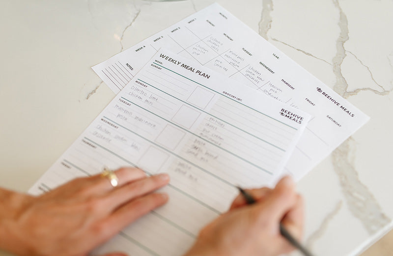 A close-up of a person's hands writing on a printed weekly meal plan sheet from Beehive Meals, placed on a light marble surface.